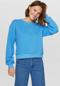 Šviesiai mėlynas džemperis su apvaliu iškirpimu, nuleistais pečiais ir raukinėtomis rankogaliais. Pora su mėlynomis džinsomis, jis turi laisvą kirpimą ir minkštą tekstūrą.