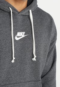 Grå hoodie i mjukt material, med en vit Nike-logotyp på bröstet och vita dragsko med tofsar.