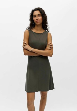 Femme aux cheveux bouclés foncés, debout, bras croisés, portant une robe sans manches vert olive longueur genou sur fond blanc uni.