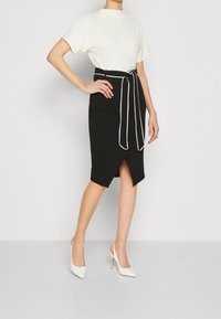 Svart pennkjol med vit kant och slipsbelte, med en hög slits. Matchad med en vit t-shirt med kort ärm och vita klackar.