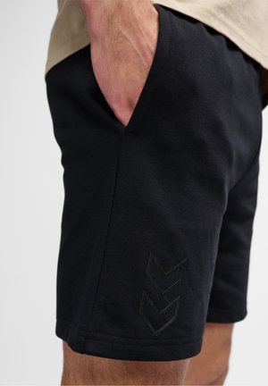 Pantalones cortos deportivos negros de textura suave, con bolsillos laterales y un sutil logo negro en forma de cheurón en la parte inferior izquierda. Diseño y ajuste sencillo.