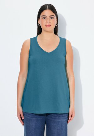 Junge Frau steht vor einer weißen Wand, trägt ein türkisfarbenes ärmelloses V-Ausschnitt-Top und blaue Jeans, schaut nach vorne mit neutralem Gesichtsausdruck.