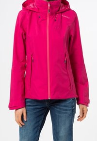 Giacca a cappuccio rosa brillante con zip frontale e tasche con zip, indossata con jeans blu, che mostra le braccia e il torso di una persona.