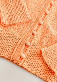 Cardigan orange texturé avec des manches côtelées et cinq boutons recouverts de tissu sur le devant. Matériau doux et duveteux sur l'ensemble.