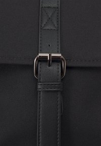 Sac messager noir avec un extérieur en tissu texturé. Doté d'une sangle en cuir noir et d'une boucle en métal argenté pour la fermeture.