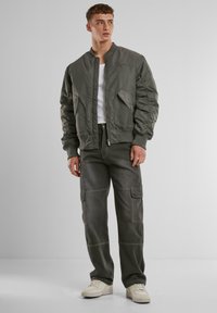 Olíva zöld bomber dzseki quilted textúrával, bordázott mandzsettákkal és két oldalsó zsebbel; cargo nadrággal és krémszínű sportcipővel párosítva.