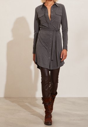 Kvinde iført en grå skjortekjole med lange ærmer og bælte, mørkebrun læderbukser og brune ruskindsstøvler, der går indendørs.