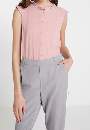 Blouse rose sans manches avec col à volants et boutons, associée à un pantalon taille haute gris clair, main du mannequin dans la poche.