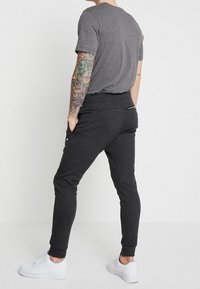 Grå bomullströja och svarta tapered sweatpants med sidofickor, elastiska muddar och vita sneakers. Tatueringar synliga på armen.