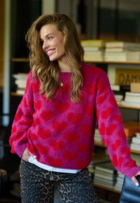 Vzor červených srdcí na jasně růžovém huňatém svetru, oversized střih s manžetami. V kombinaci s džínami s leopardím vzorem. Teplá, strukturovaná tkanina.