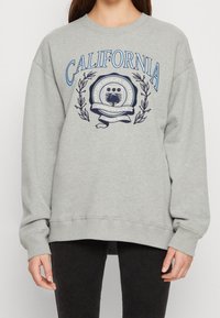 Person som bär en grå sweatshirt med blå texten "CALIFORNIA" och ett emblem med "East Los Angeles Athletics Commons Club".