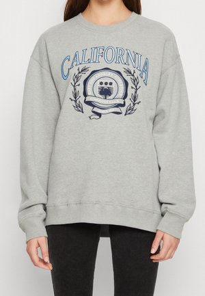 Personne portant un sweat-shirt gris avec le texte bleu "CALIFORNIA" et un blason comportant l'emblème du "East Los Angeles Athletics Commons Club".