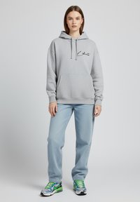 Felpa grigia con cappuccio e tasca frontale, con logo stampato nero. Abbinata a jeans azzurri a vestibilità comoda e scarpe da ginnastica colorate.