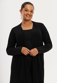 Lange zwarte cardigan met lange mouwen, voorzien van gouden studs op de manchetten. Gemaakt van zachte stof met een gedrapeerd ontwerp.