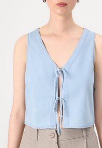 Lichtblauwe mouwloze crop top van zachte denimstof, met een diepe V-hals en een sluiting aan de voorkant met twee hangende strikjes.