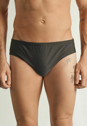 Męski tors w ciemnych kąpielówkach z logo „IUMAN”, pokazujący tatuaże dłoni, anatomicznego serca i tekstu na udzie i ramieniu.