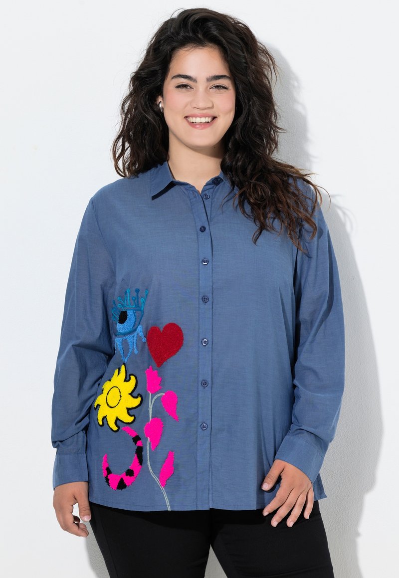 Camicia blu con bottoni caratterizzata da vivaci ricami floreali e astratti nei toni di giallo, rosa e turchese su un lato. Maniche lunghe.