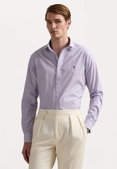Camisa de vestir a rayas en un ligero tono púrpura, con cuello clásico y cierre de botones, que presenta un pequeño logotipo. Combinada con pantalones plisados de color crema.