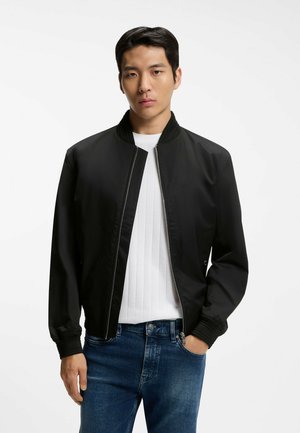 Ung mand med kort mørkt hår iført en sort lynlåstrøje over en hvid ribbet skjorte og blå jeans, stående mod en hvid baggrund.