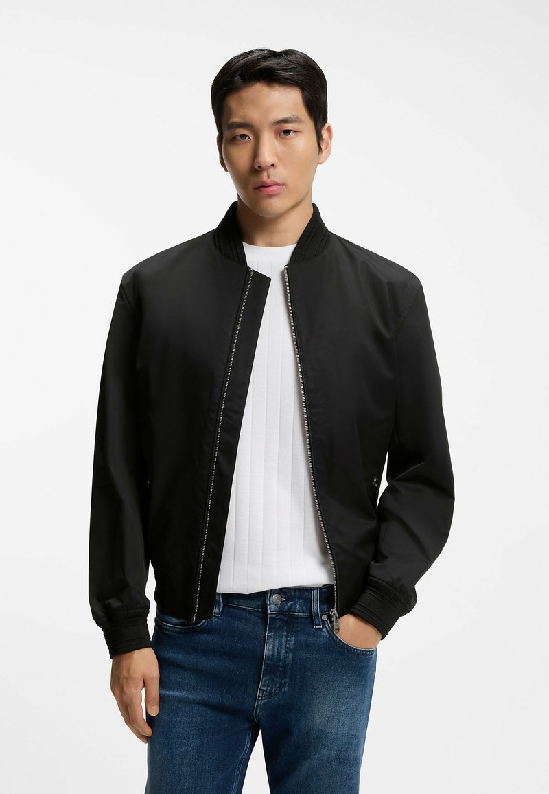 Młody mężczyzna o krótkich ciemnych włosach, ubrany w czarną kurtkę na zamek błyskawiczny na białą prążkowaną koszulkę i niebieskie dżinsy, stojący na białym tle.