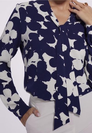 Blouse bleu marine avec un motif floral blanc, manches longues et décolleté en V. Dotée d'un lien au niveau du col et d'un tissu lisse et léger.