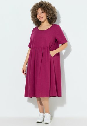 Donna con capelli ricci che indossa un vestito largo magenta fino al ginocchio e scarpe da ginnastica bianche, in piedi con una mano in tasca.