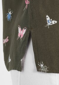 Grüner Stoff mit einem seitlichen Schlitz, verziert mit pinken und blauen Schmetterlings- und Käferdruckmotiven, getragen über weißen, strukturierten Hosen.