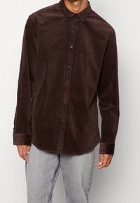 Hnědá manšestrová košile s límečkem na knoflíky, předními knoflíky a dlouhými rukávy, vyznačující se texturovanou látkou a volným střihem.