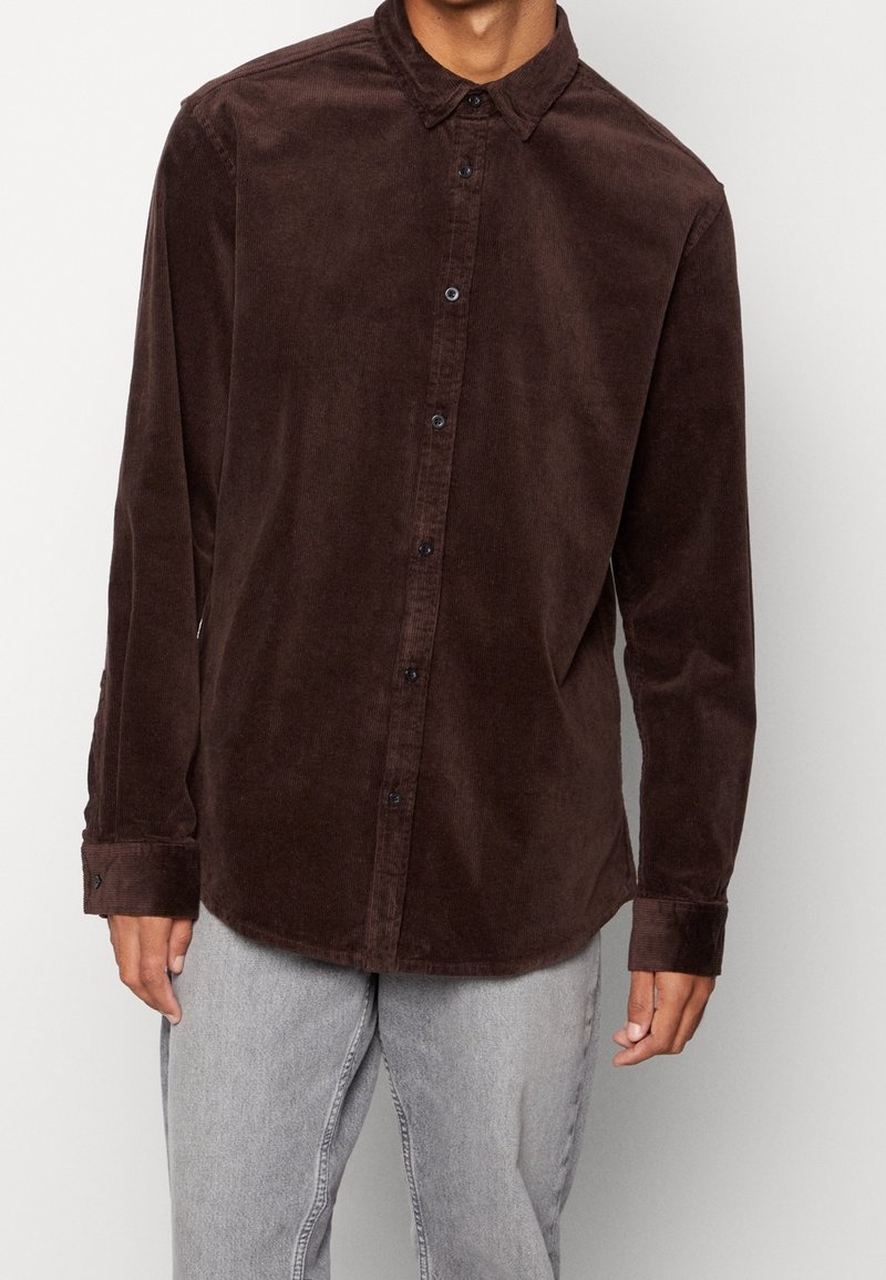 Hnědá manšestrová košile s límečkem na knoflíky, předními knoflíky a dlouhými rukávy, vyznačující se texturovanou látkou a volným střihem.