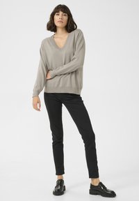 Beige V-Ausschnitt-Pullover mit langen Ärmeln, lockerer Schnitt, kombiniert mit schwarzen Slim-Fit-Hosen und schwarzen Slippern, strukturierter Strickstoff.