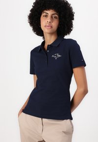Marineblå polotrøye i bomullsblanding, med krage som kan knytes opp og brodert logo-design på brystet. Fitted fasong med korte ermer.