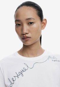 Valge puuvillane T-särk, millel on musta tikandiga tekst "desigual" ja "proud to be different". Särgil on ümar kaelus ja lühikesed varrukad.