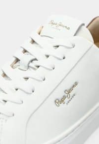 Sneakers bianchi in pelle con superficie testurizzata, lacci bianchi piatti e un logo dorato sul lato. Presentano un interno imbottito color tan.