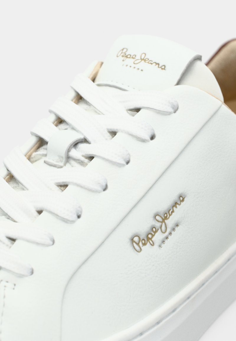 Baskets en cuir blanc avec une surface texturée, lacets blancs plats et un logo doré sur le côté. Elles possèdent un intérieur rembourré de couleur beige.