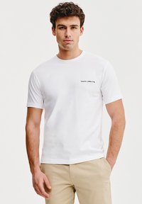 Biała koszulka z krótkim rękawem, okrągłym dekoltem i małym czarnym logo na lewej piersi, wykonana z gładkiej bawełnianej tkaniny. Zaprojektowana do codziennego noszenia.