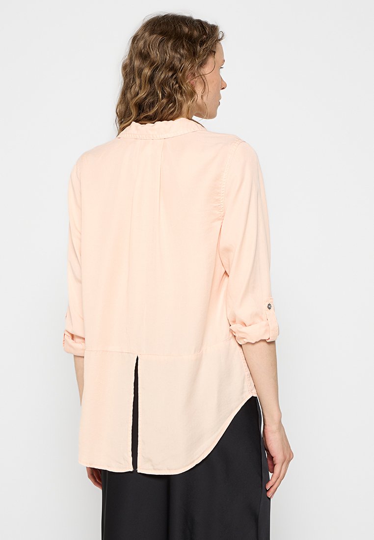 Camisa de melocotón claro con mangas largas, puños enrollables, dobladillo redondeado y un pliegue central en la parte posterior para mayor transpirabilidad. Textura de tela suave.