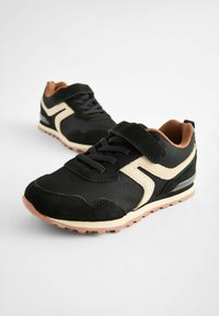 Chaussures de sport noires en mesh et daim, avec une bande diagonale crème, semelles en caoutchouc texturées et bride velcro.