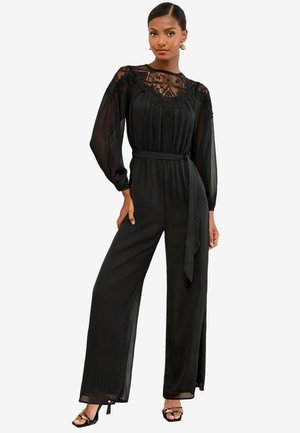 Woman wearing a black lace and chiffon long-sleeve jumpsuit with a waist tie and wide-leg pants, paired with black open-toe heels.