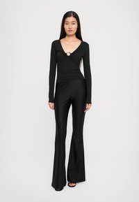 Black long-sleeve top with a twisted design and circular gold accent, paired with flared black pants made of a smooth, shiny material.