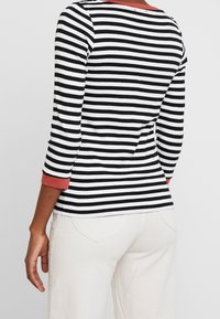 Camicia a maniche lunghe a righe in bianco e nero con polsini di colore corallo. Tessuto morbido con una silhouette aderente, caratterizzata da un dettaglio di cucitura sul retro.