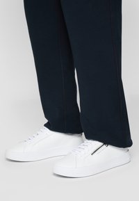 Hvite sneakers med glatt skinnfinish, med glidelås på siden, flate lisser og strukturert gummisåle, kombinert med mørke bukser.