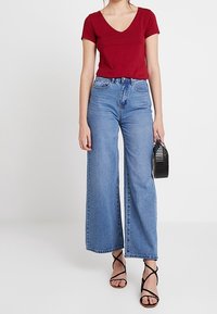 Red v-neck top paired with high-waisted, wide-leg blue jeans. Model holding a black handbag and wearing black strappy sandals.