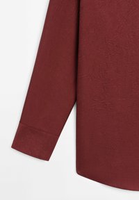 Chemise à manches longues en tissu rouge foncé texturé avec un poignet à un seul bouton et un ourlet arrondi sur fond blanc.
