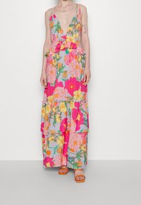 Mujer vistiendo un vestido largo sin mangas de flores con escote en V profundo y volantes, combinado con sandalias abiertas de color naranja, de pie frente a un fondo sencillo.