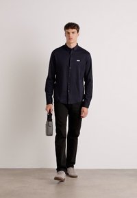 Marineblå skjorte med knapper og en liten logo, kombinert med sorte bukser og grå sko. Personen holder en kompakt grå bag. Glatt stofftekstur.
