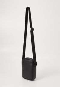 Petit sac en cuir noir porté en bandoulière, avec une sangle réglable et une fermeture éclair, isolé sur un fond blanc.