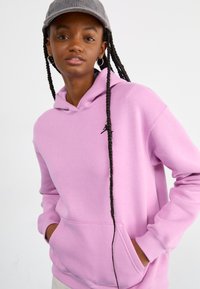 Lilac hooded sweatshirt made of soft fabric, featuring a kangaroo pocket and a small black logo on the chest. Long braids visible.
