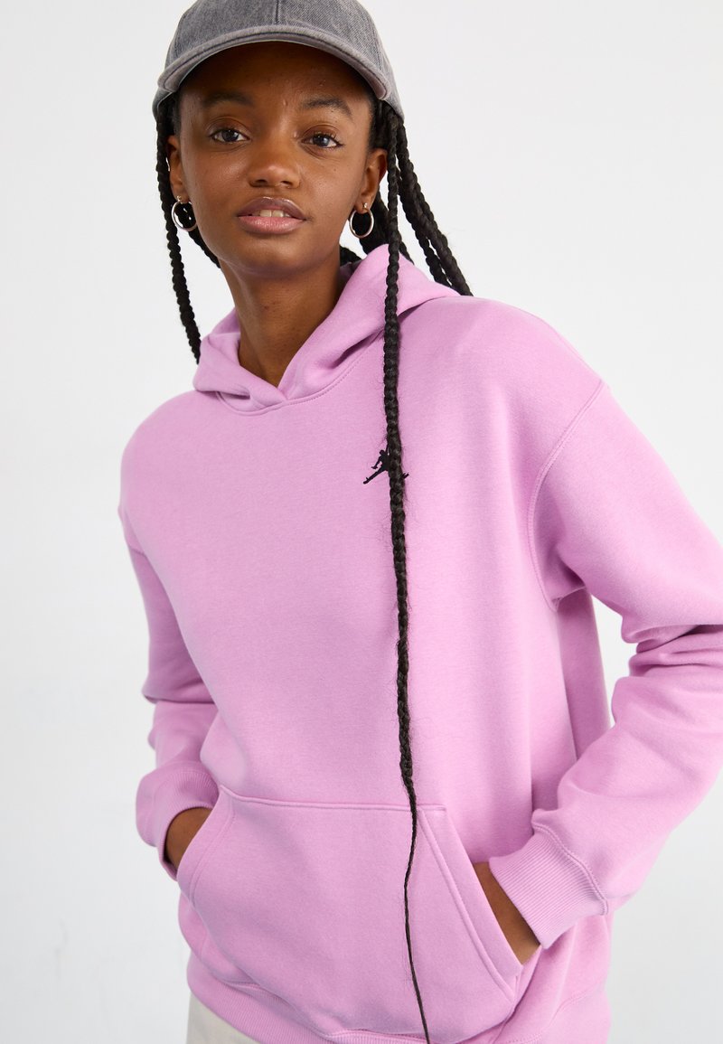 Fioletowy sweatshirt z kapturem wykonany z miękkiego materiału, z kieszenią kangurką i małym czarnym logo na klatce piersiowej. Widoczne długie warkocze.