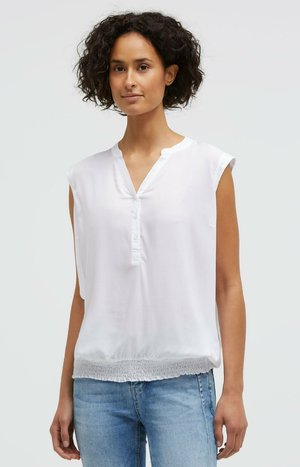 Woman with short curly hair wearing a white sleeveless blouse with buttoned V-neck and blue jeans, standing against a plain background.