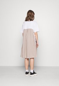 Valkoinen lyhytnhelmainen toppi, jossa on beige raidallinen hame, jossa on napit sivussa ja laskostettu muotoilu, esiteltynä takaa.